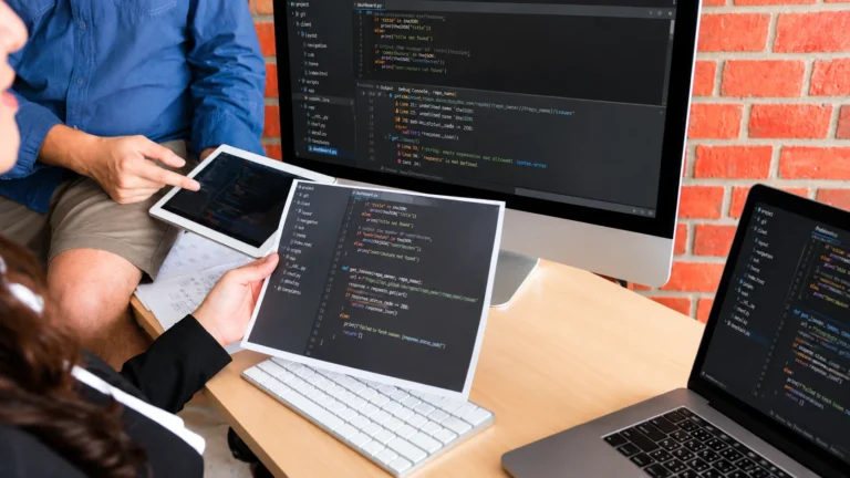 Team of developers collaborating on a website redesign project using multiple screens displaying code, including a desktop monitor, laptops, and a tablet in a modern workspace.